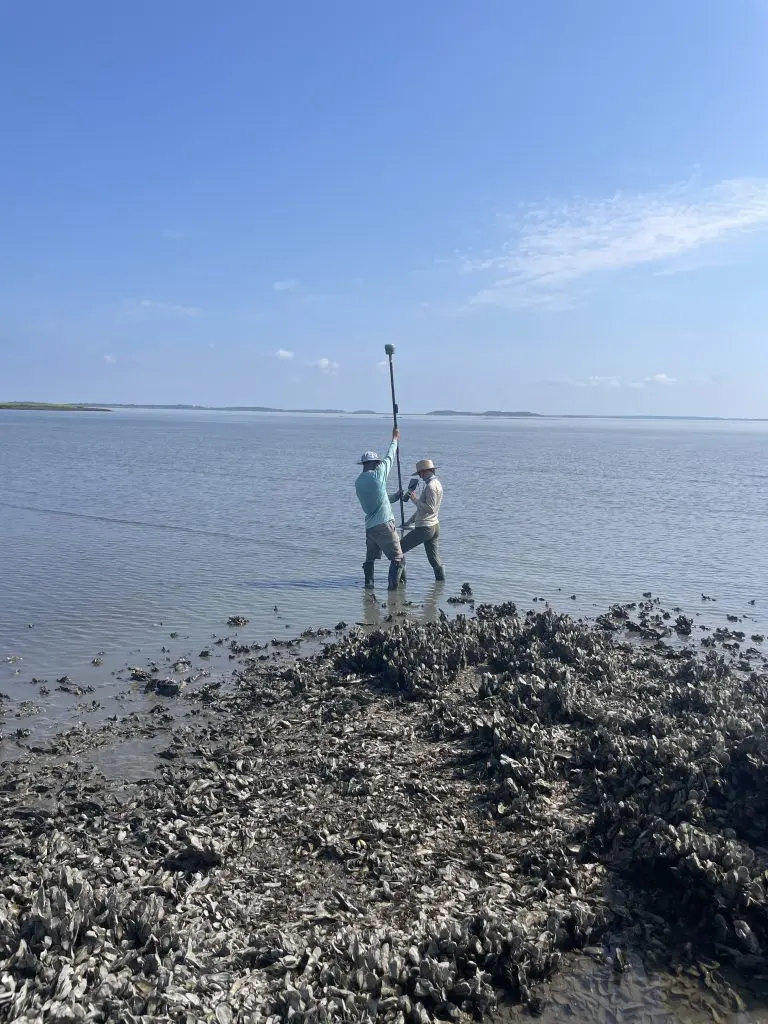 Two people in the water holding a GPS unit on a pole with oysters in the foreground