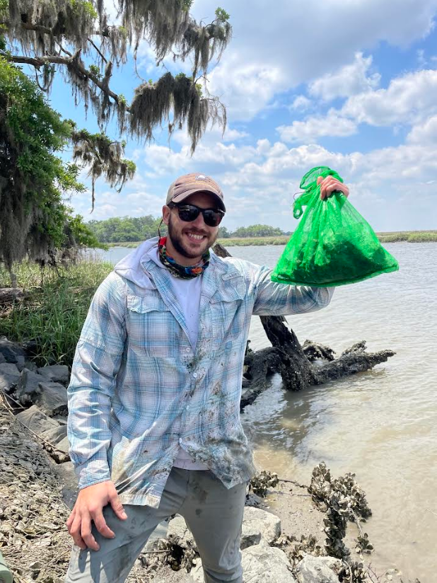 A young man holding a a mesh bag of oysters on the edge of a tidal creek.